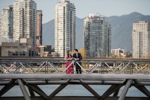 Two figures in formal attire stand on a metal bridge with modern high-rise buildings and mountains in the background.