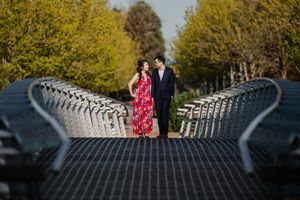 A couple stands on a bridge with metal railings creating leading lines while autumn trees line the background.