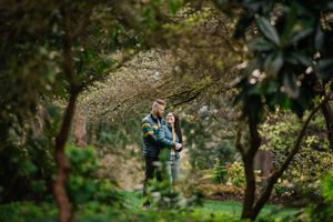 Romantic couple embraces in a lush green garden setting framed by branches and sunlit foliage.