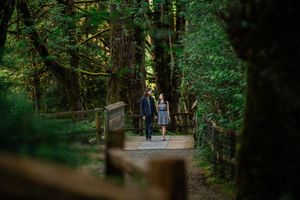 Two people stroll along a wooden boardwalk path through a serene, lush green forest.