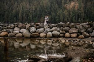 A couple stands together on large river rocks against a backdrop of evergreen forest and calm waters.