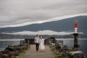 A pair walks along a rocky jetty with a lighthouse and misty mountains in the background.