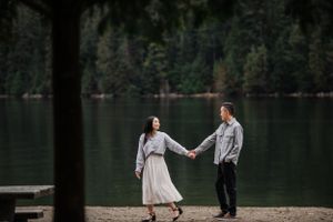 Two people hold hands by a serene lake surrounded by dense forest.