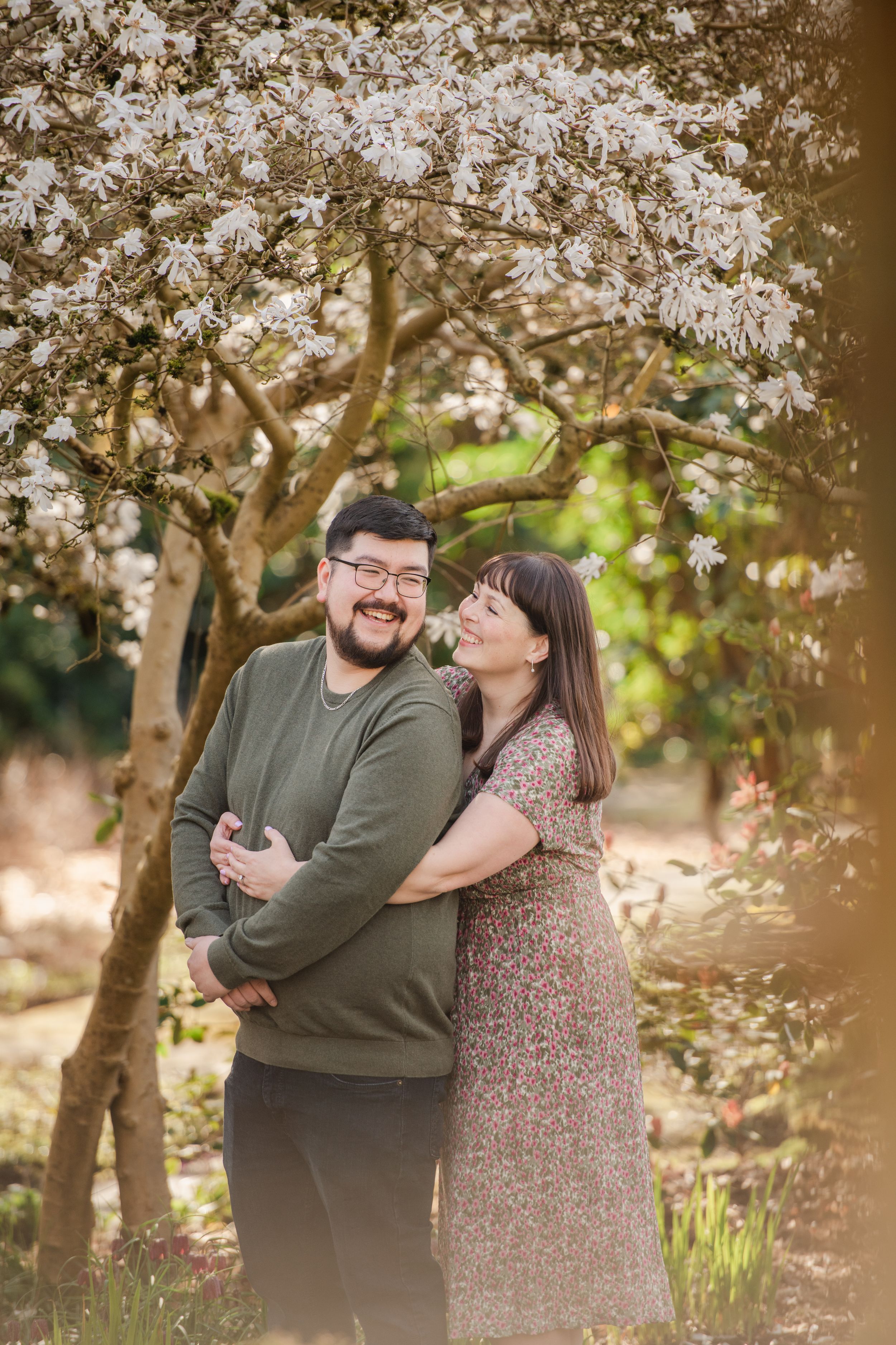 A couple embraces and smiles under a blooming white magnolia tree in a lush garden during golden hour.