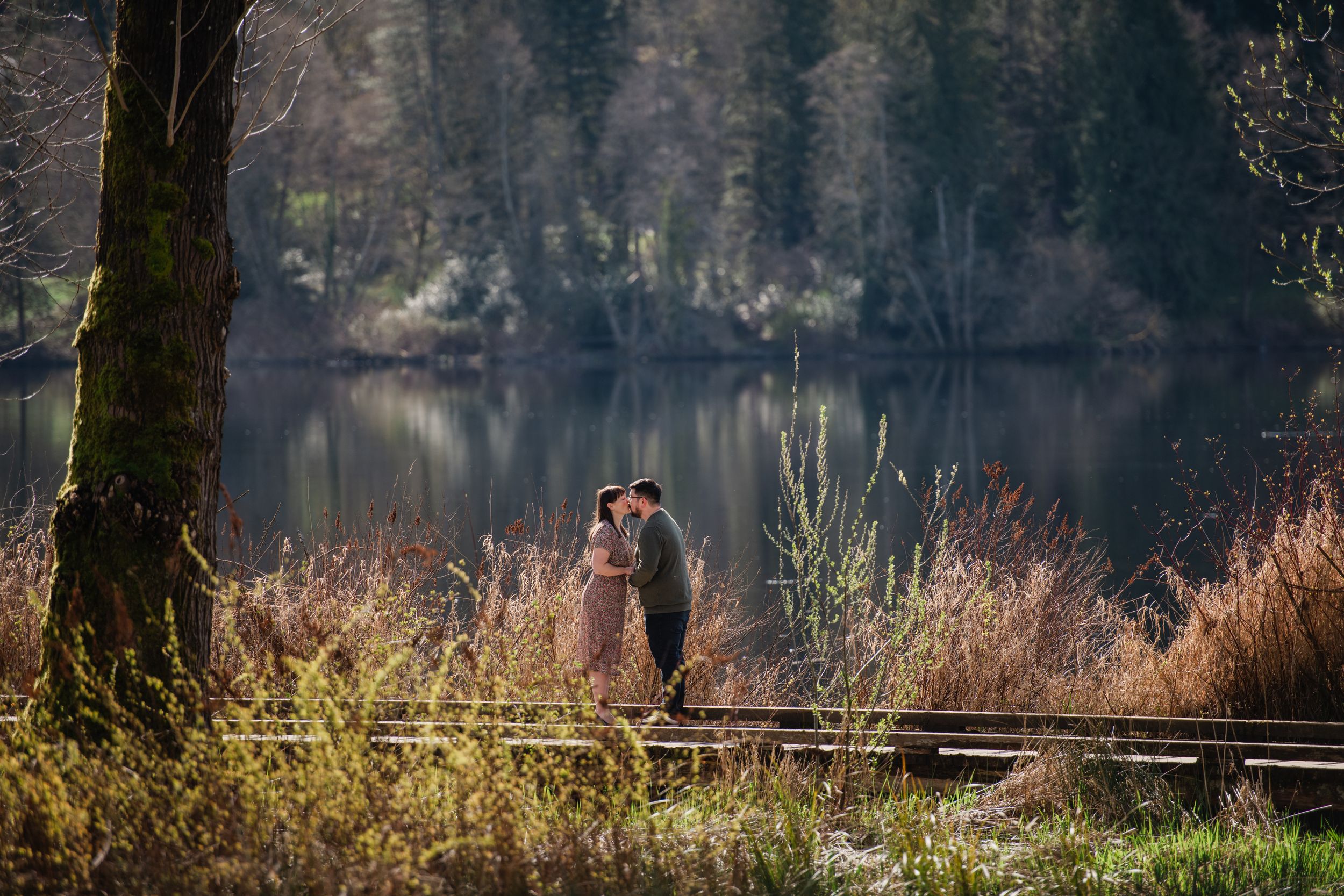 Couple embracing near a serene lake surrounded by autumn trees and golden shrubs in a romantic outdoor setting.