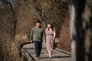 Couple holding hands walking on a wooden boardwalk path surrounded by autumn trees and tall grass.