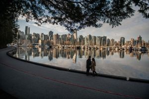 Two people walking along Vancouver's seawall at Stanley Park with downtown skyline reflecting in calm harbor waters at sunset.