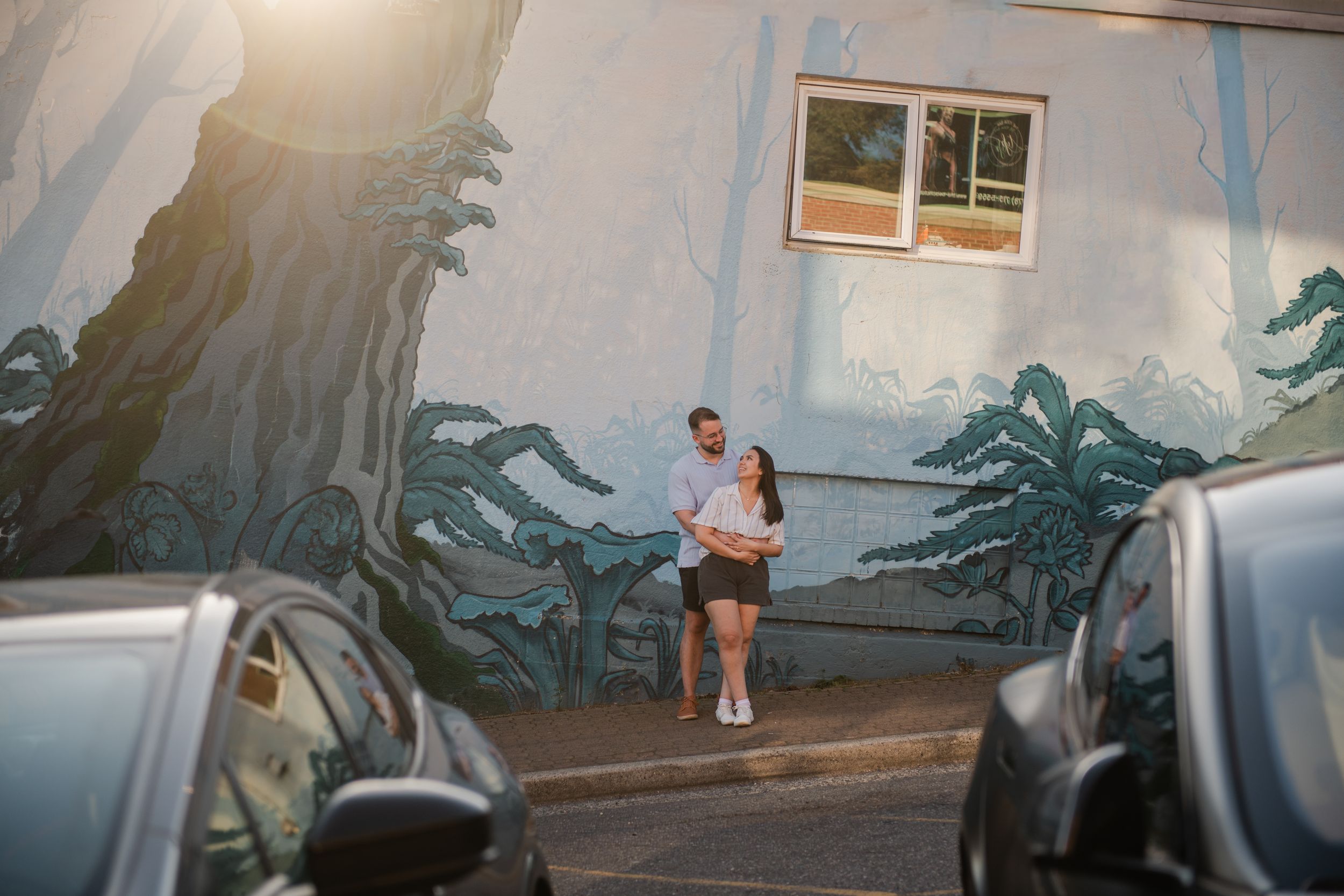 Couple embracing in front of a colorful nature mural on a building wall, with parked cars in the foreground.