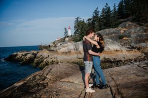 A romantic moment shared between a couple standing on rocky coastal cliffs overlooking the ocean with evergreen trees.