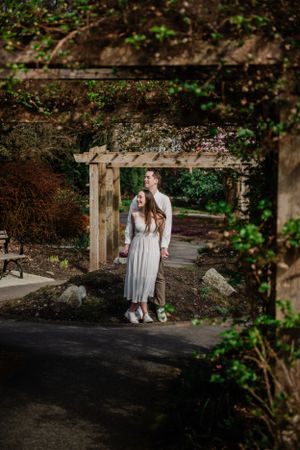 A couple stands beneath a rustic wooden arbor on a garden path surrounded by lush greenery and natural light.