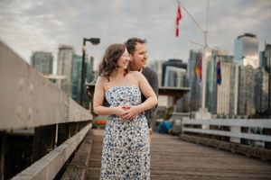 Couple embraces on wooden pier with downtown Vancouver skyline and Canadian flag in background on summer evening.