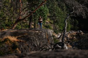 A couple embraces on a rugged cliff edge surrounded by towering pine trees in a dramatic outdoor setting.
