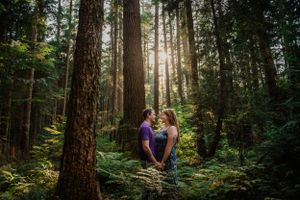 Two people share an intimate moment in a sunlit forest surrounded by tall evergreen trees.