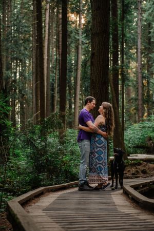 A couple embraces on a wooden boardwalk trail through a dense forest with their black dog.