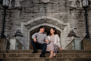 Couple sits on stone steps in front of grand gothic archway during romantic engagement photo session.