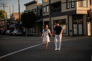 A couple holding hands walks across an urban street intersection at sunset with storefronts and buildings in the background.