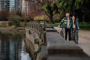 Two people walking with a stroller along a waterfront pathway beside a stone wall in an urban park.