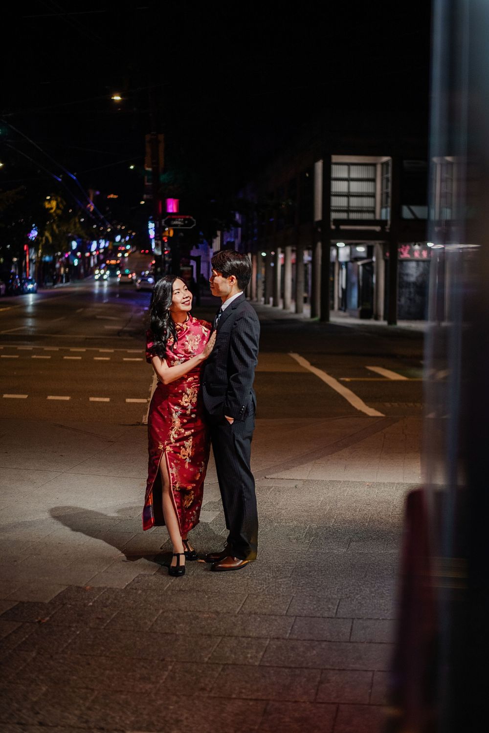 Couple in formal wear embracing on a city sidewalk at night, woman in red qipao dress, man in dark suit.