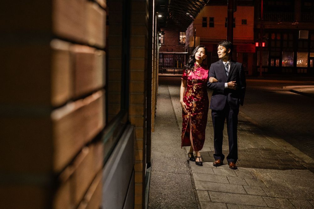 Couple in elegant attire, woman in red qipao and man in suit, standing on a city sidewalk at night.
