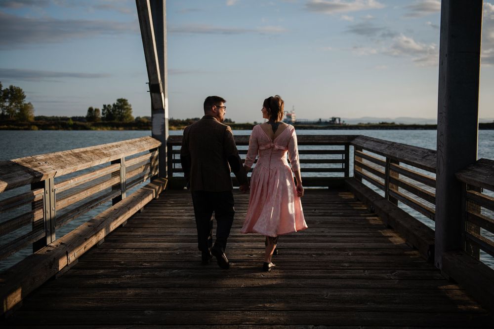Couple walking hand-in-hand on a wooden pier at sunset, woman in pink dress, man in dark suit, waterfront view.