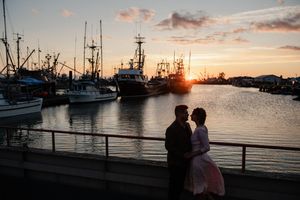 Couple silhouetted at sunset by a marina with fishing boats, sharing a romantic moment on a waterfront dock.
