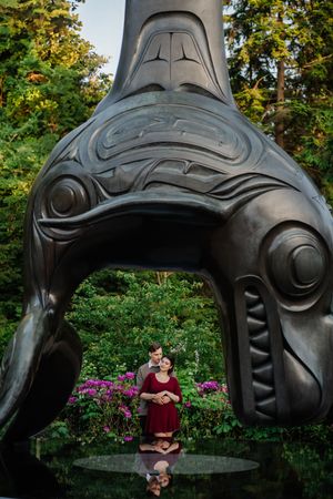 Large carved totem sculpture in the style of Pacific Northwest Coast art stands in a garden with purple flowers.