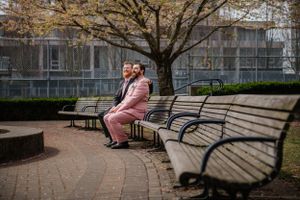 A couple sits together on a curved park bench under a bare tree on a brick pathway.