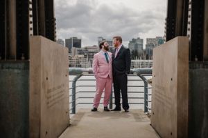 A couple in contrasting formal attire poses on an urban bridge with city skyline views.