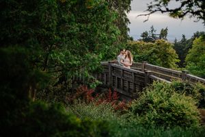 Two people stand on a scenic wooden footbridge surrounded by lush gardens and evergreen trees in a parklike setting.