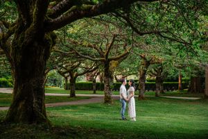 Two figures stand together under majestic old trees in a lush green park setting during golden hour.