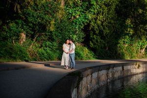 A couple embraces on a curved pathway beside a stone wall, surrounded by lush green foliage in evening light.