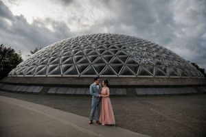A couple poses in front of a geodesic dome structure under dramatic cloudy skies.
