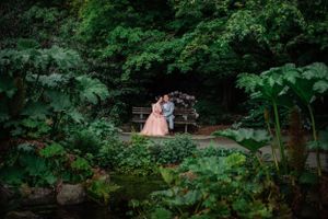 A couple stands on a bridge in a lush garden setting with green foliage and water features.