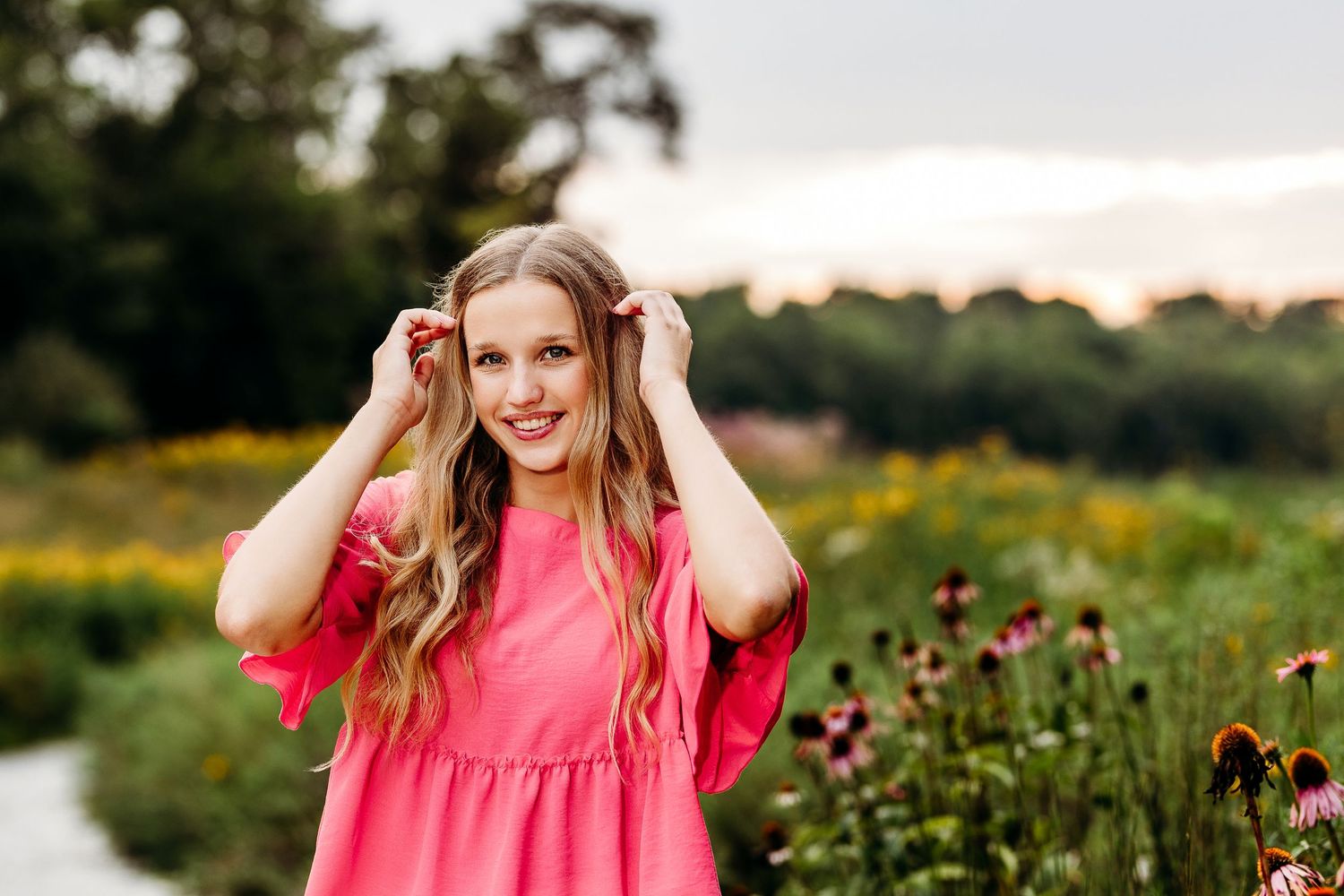 A person in pink adjusts their hair while standing in a garden of wildflowers at sunset.