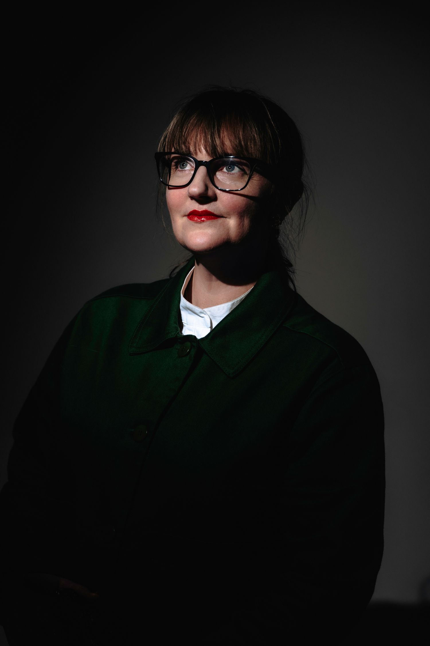 A studio portrait of photographer Rebecca McMillan wearing a green jacket, black rimmed glasses, and with red lipstick