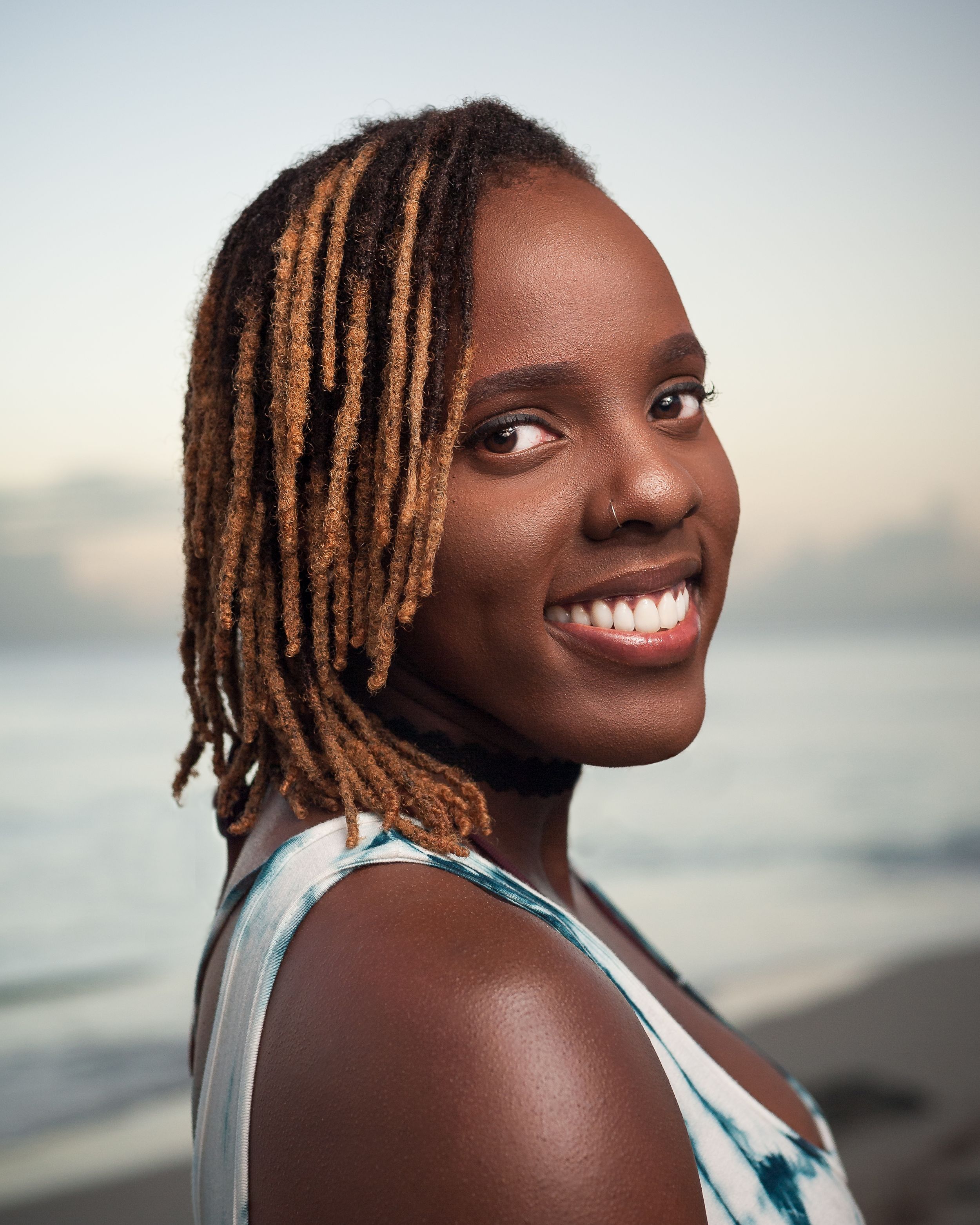 A joyful portrait in a beach setting with blonde dreadlocks and a bright smile against a blurred ocean backdrop.