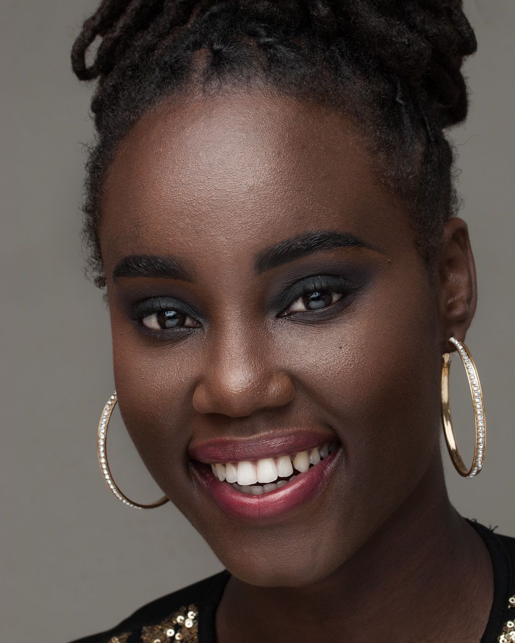 Portrait of someone in black outfit wearing gold hoop earrings and red lipstick against grey background.