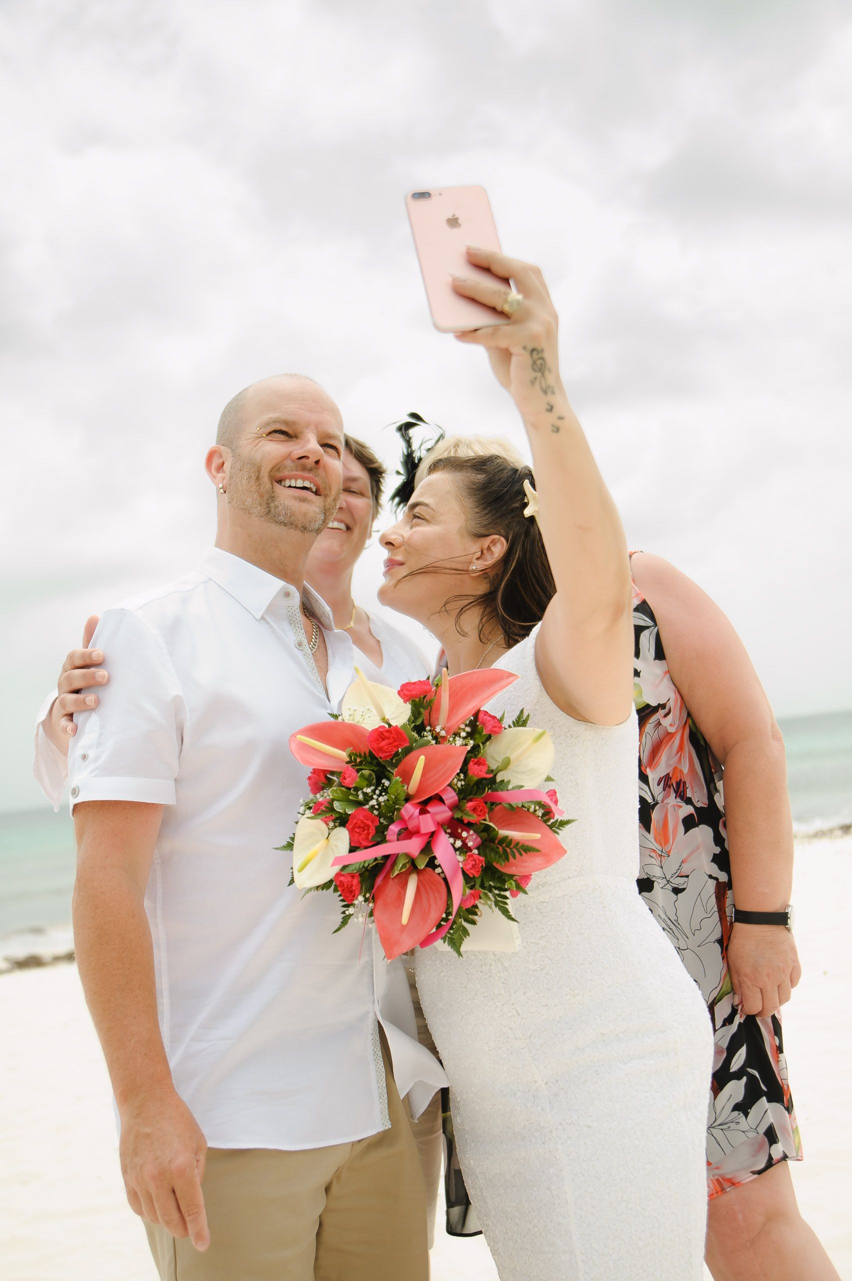 Happy couple takes selfie on beach while holding bright tropical flower bouquet during destination wedding celebration.