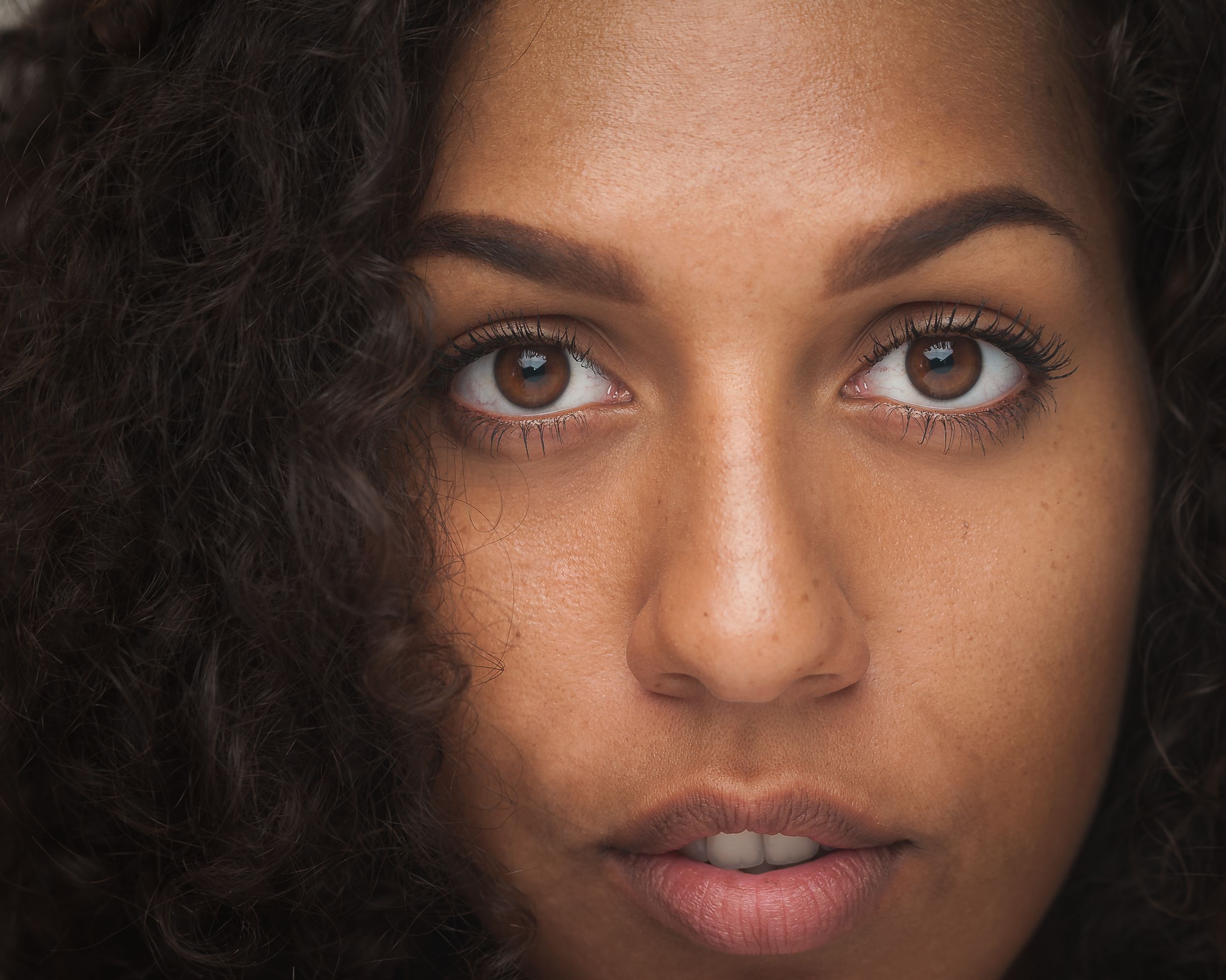 Portrait of a person with natural curly hair and warm brown eyes looking directly at camera against dark background.