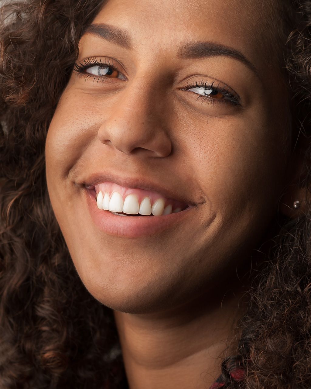 Close up portrait of a smiling person with curly hair and bright white teeth against dark background.