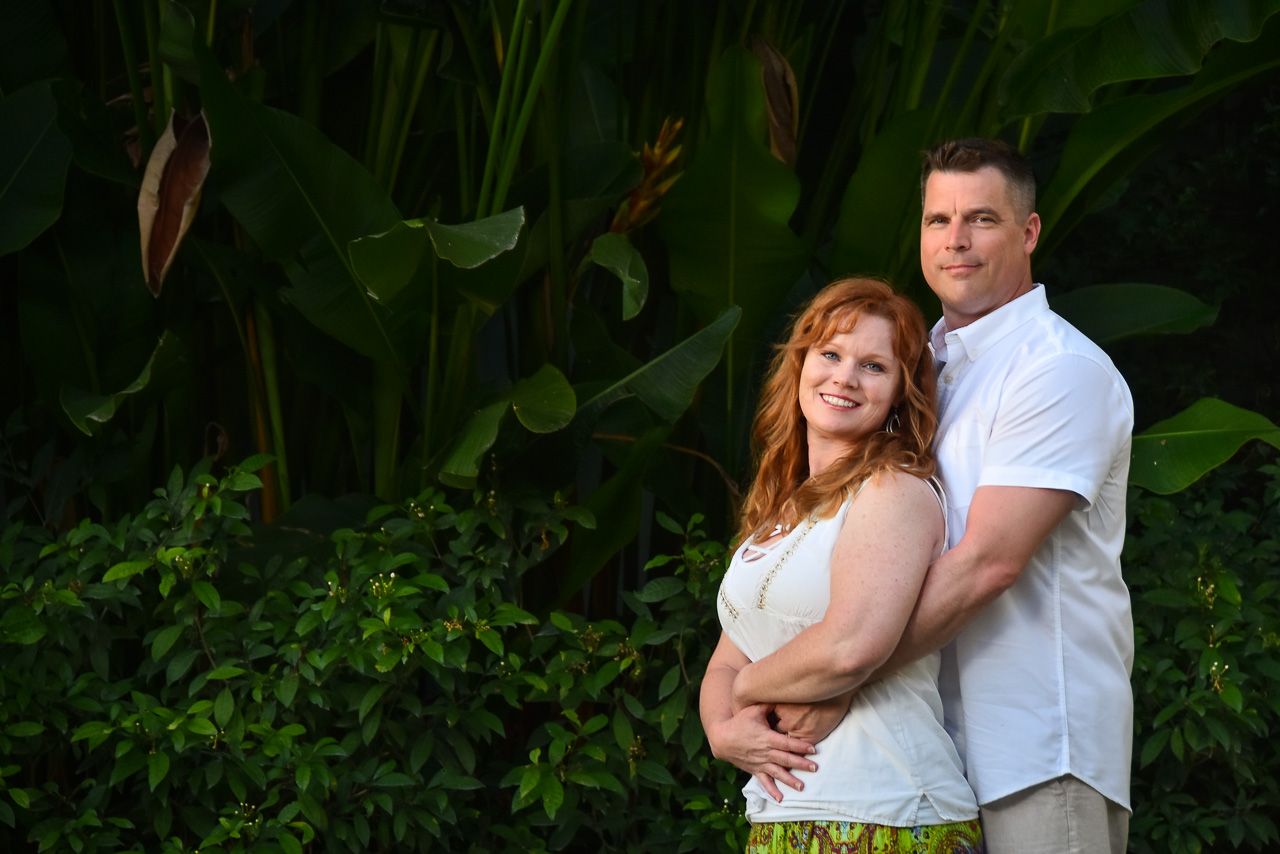 A couple poses together against lush tropical foliage in an outdoor portrait setting.
