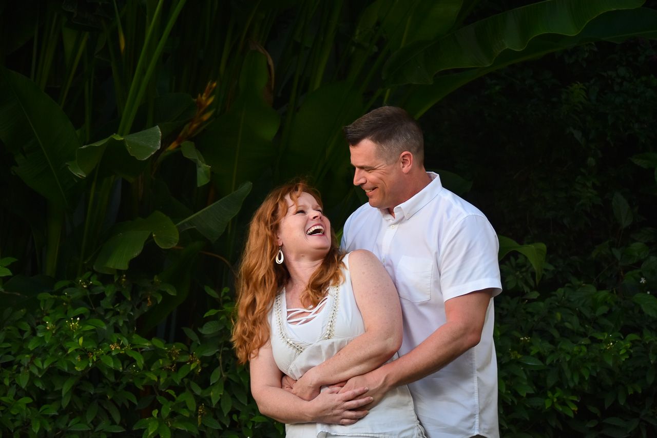 A couple shares a joyful moment together while embracing in front of lush green foliage.
