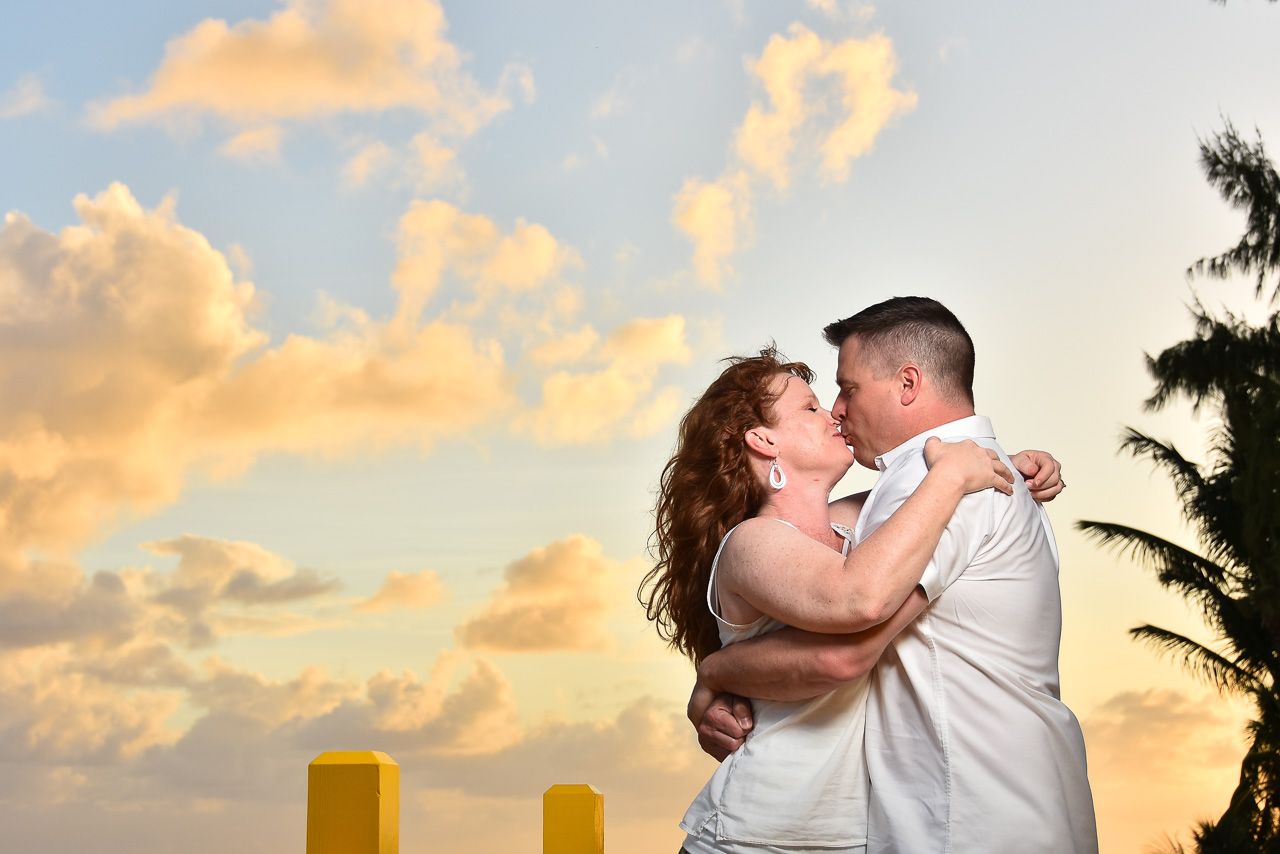 A romantic couple embraces during a warm sunset with palm trees and yellow pillars silhouetted against golden clouds.