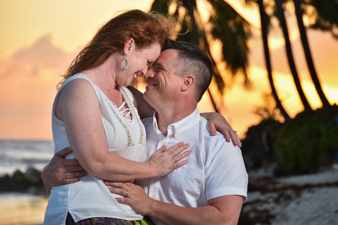 A romantic couple embraces and laughs together on a tropical beach at sunset with palm trees silhouetted in the background.