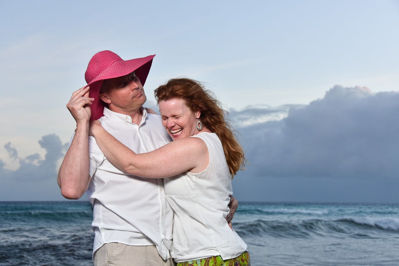 A joyful couple in white embraces while dancing together on a beach at dusk with stormy clouds in the background.