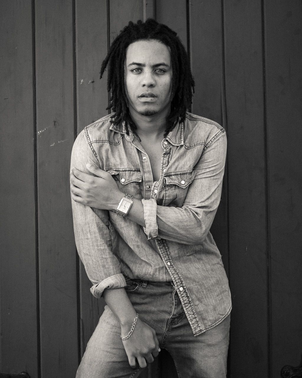 Black and white artistic portrait of a person with dreadlocks wearing a denim shirt posed against a textured wall.