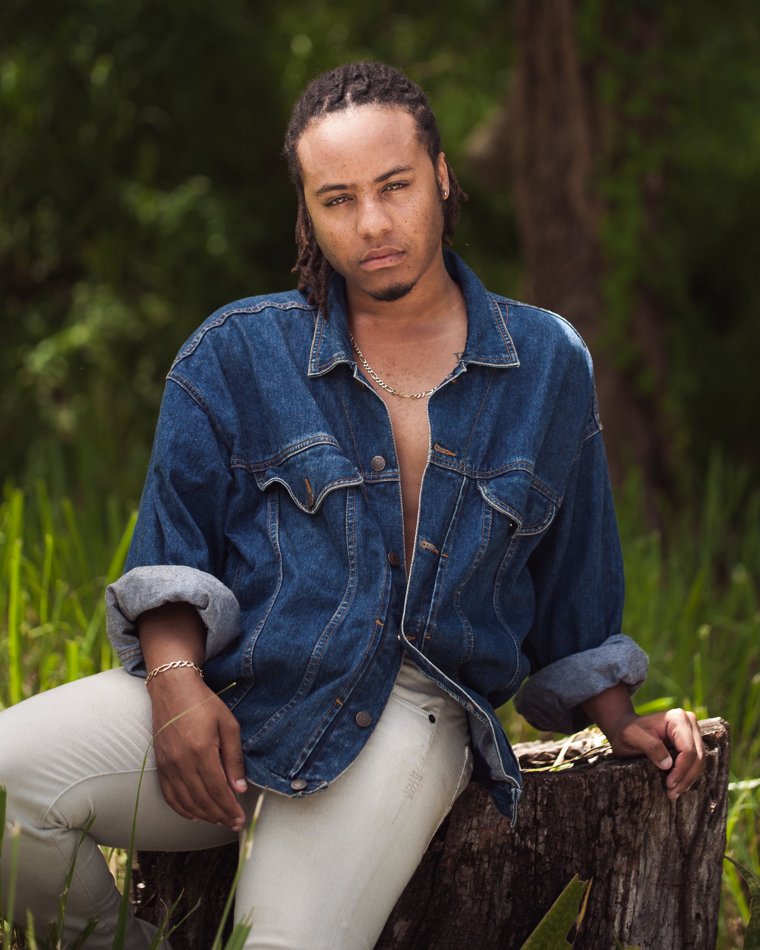 Person in casual denim jacket and light pants sits on rustic tree stump outdoors against green natural background.