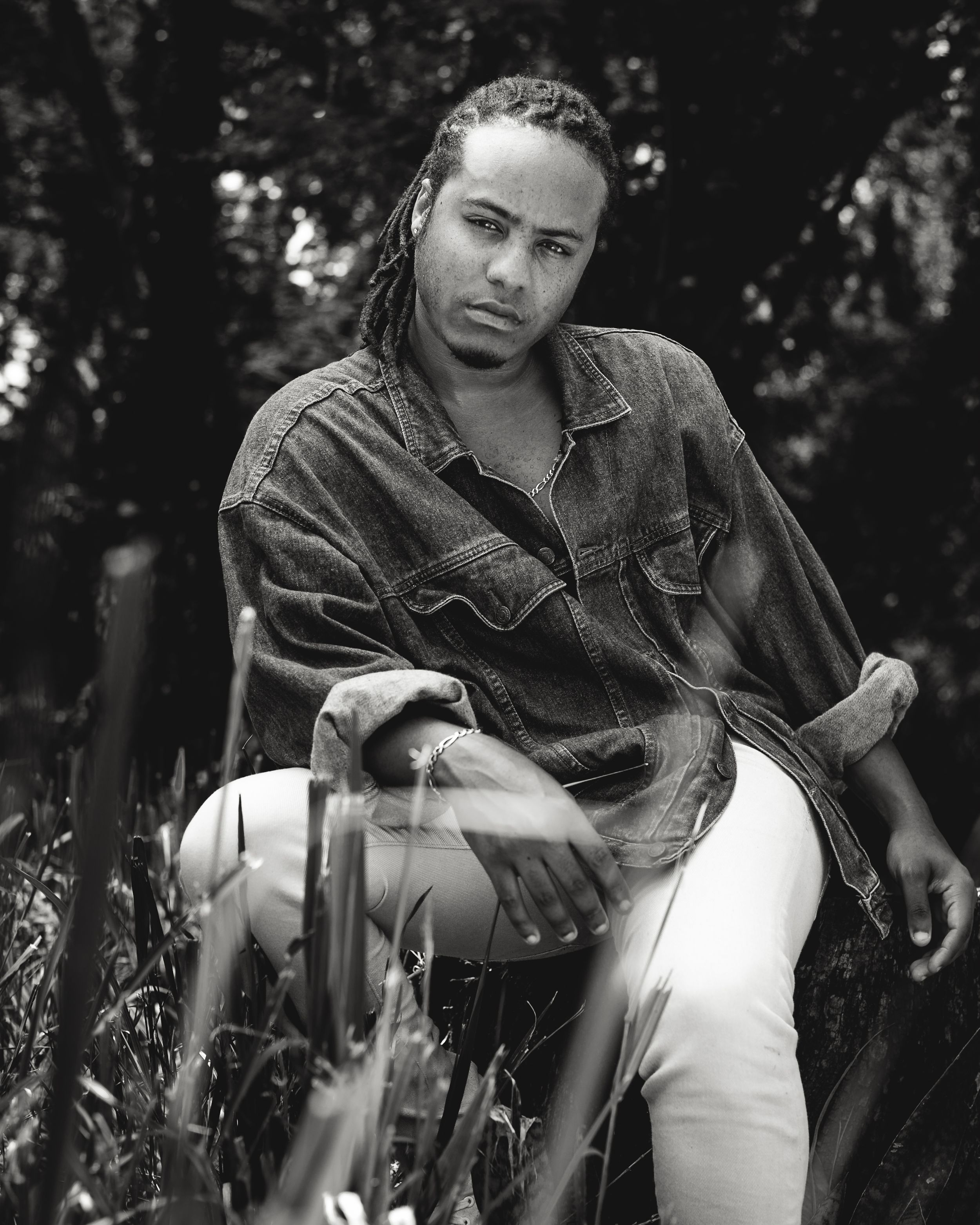 Black and white portrait photo showing person in denim jacket and white pants sitting outdoors in a natural setting.