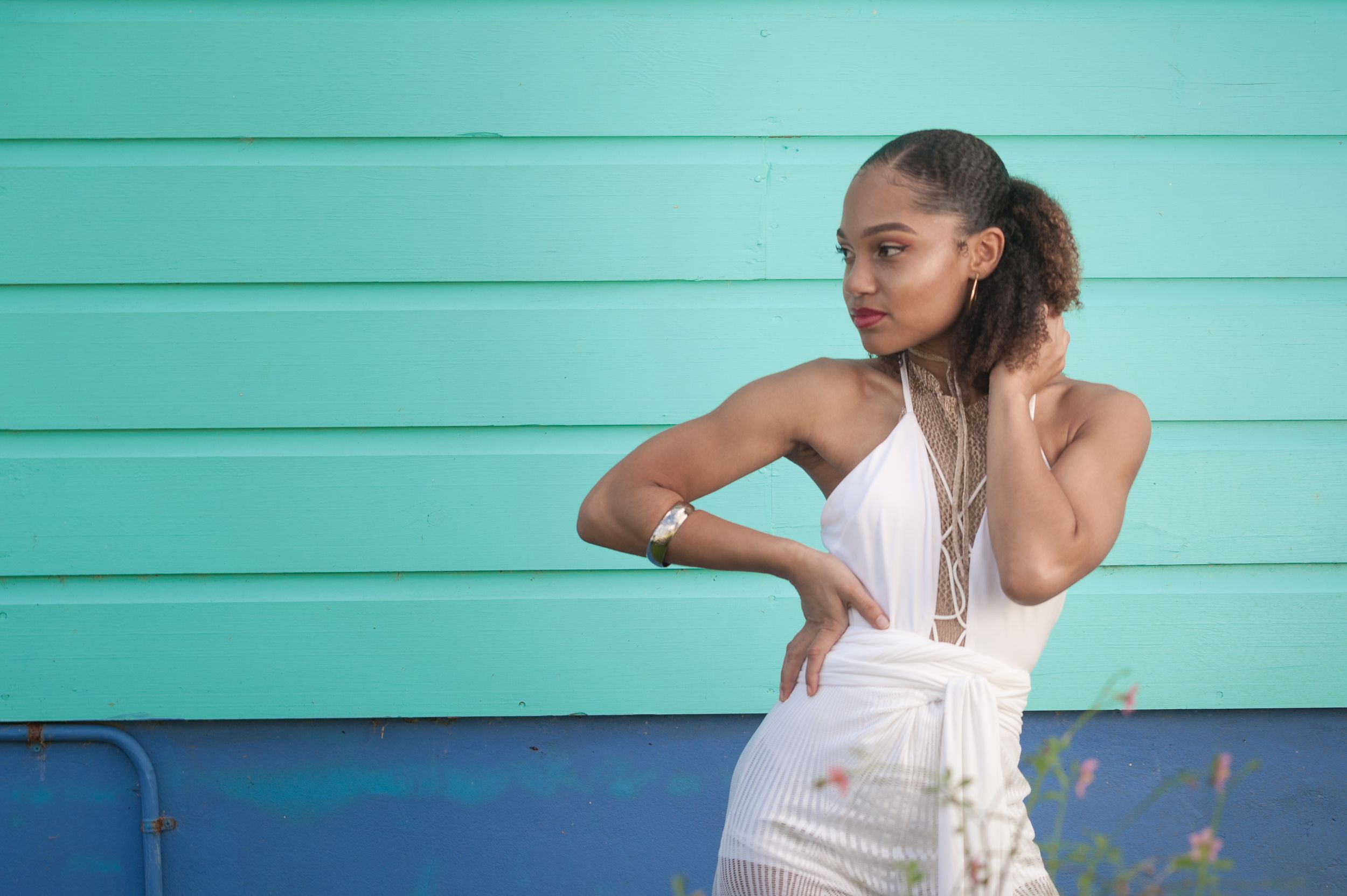 Fashion model in white dress poses elegantly against turquoise wall during outdoor photoshoot.