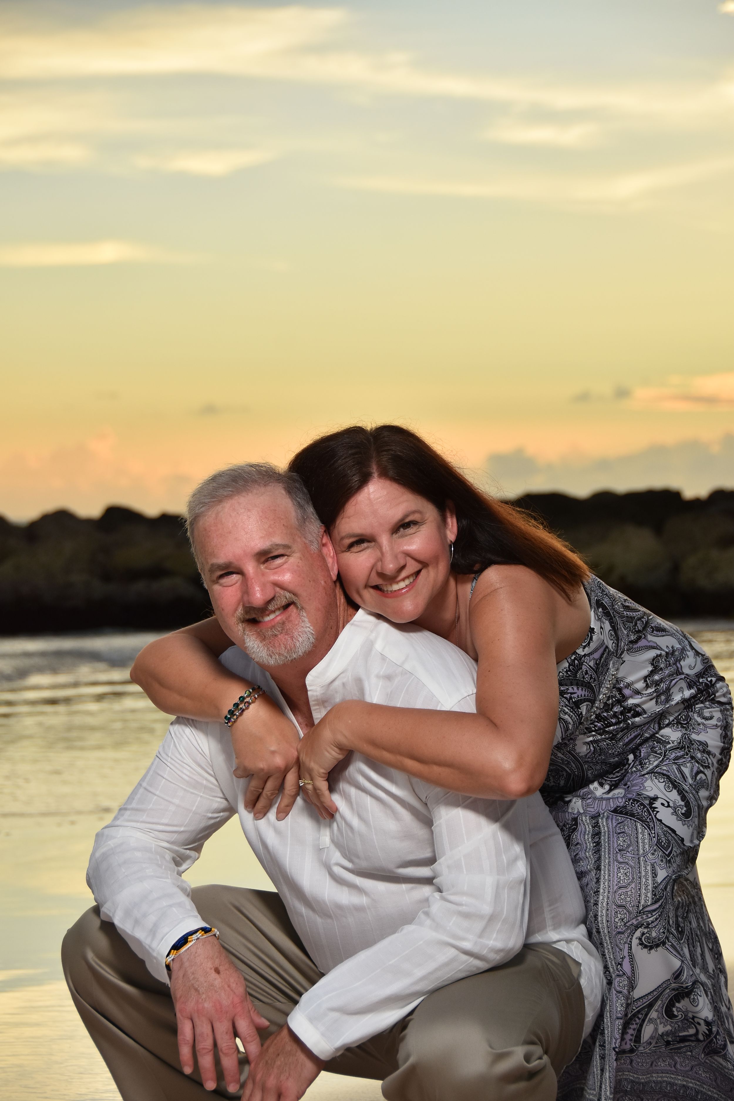 A couple shares an affectionate moment together on the beach at sunset with water and rocks in the background.